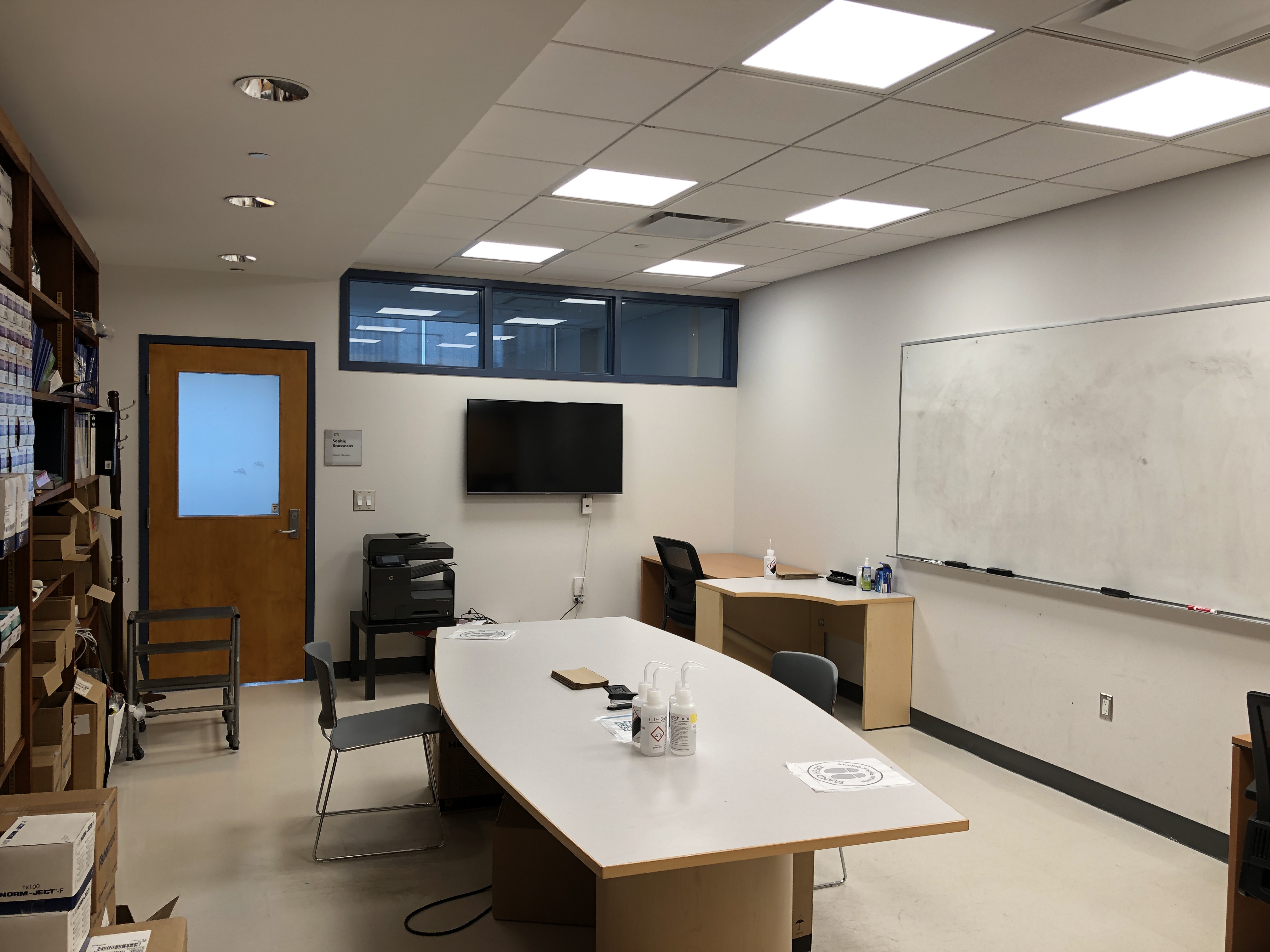 Photo of the communal group room used for meetings, eating meals, and socializing. Bookcases on the left, big oval table in the middle, and a big whiteboard on the wall on the right side.