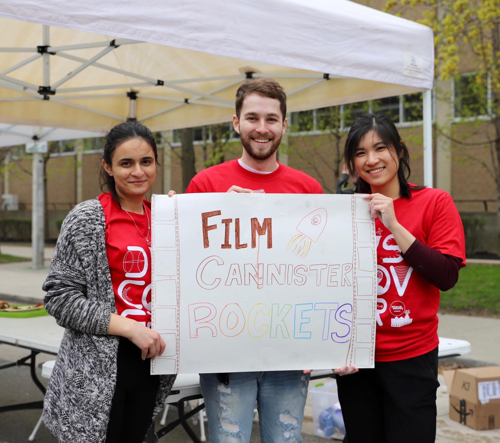 Medina, Mike, and Emily wearing red t-shirts and holding up a sign reading "film cannister rockets" for the Science Rendezvous annual event.