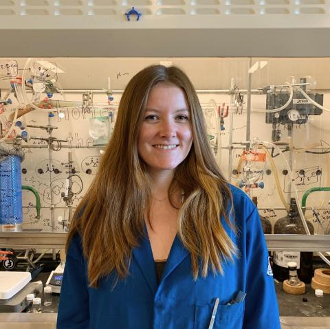 Katie in a blue lab coat standing in front of a lab fume hood.