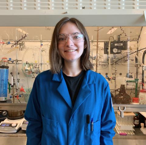 Sam in a blue lab coat standing in front of a lab fume hood.