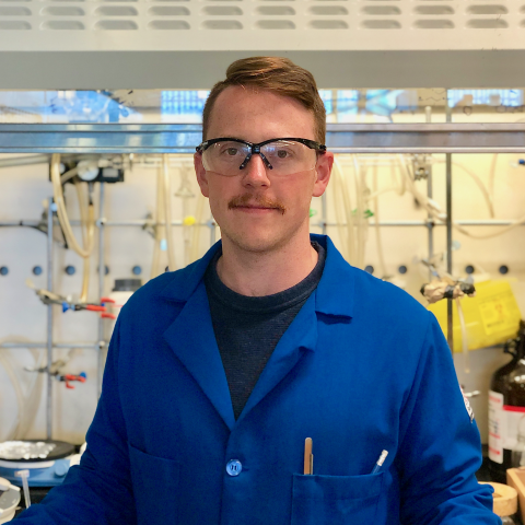 James in a blue lab coat and safety goggles standing in front of a lab fumehood