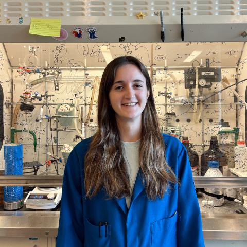 Livi in a blue lab coat standing in front of a lab fume hood.