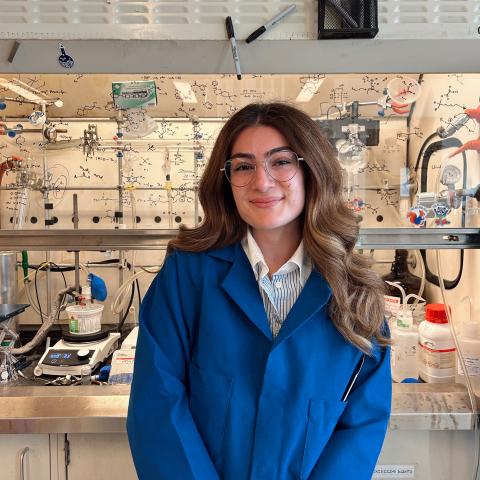 Gabriella in a blue lab coat standing in front of a lab fume hood.