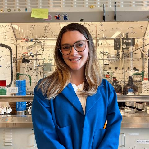 Marissa in a blue lab coat standing in front of a lab fume hood.
