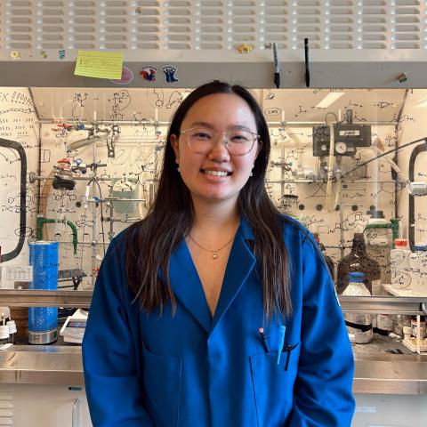 Julie in a blue lab coat standing in front of a lab fume hood.