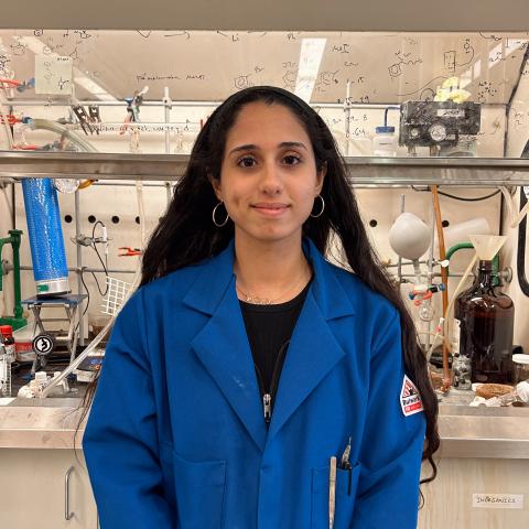 Negin in a blue lab coat standing in front of a lab fume hood.